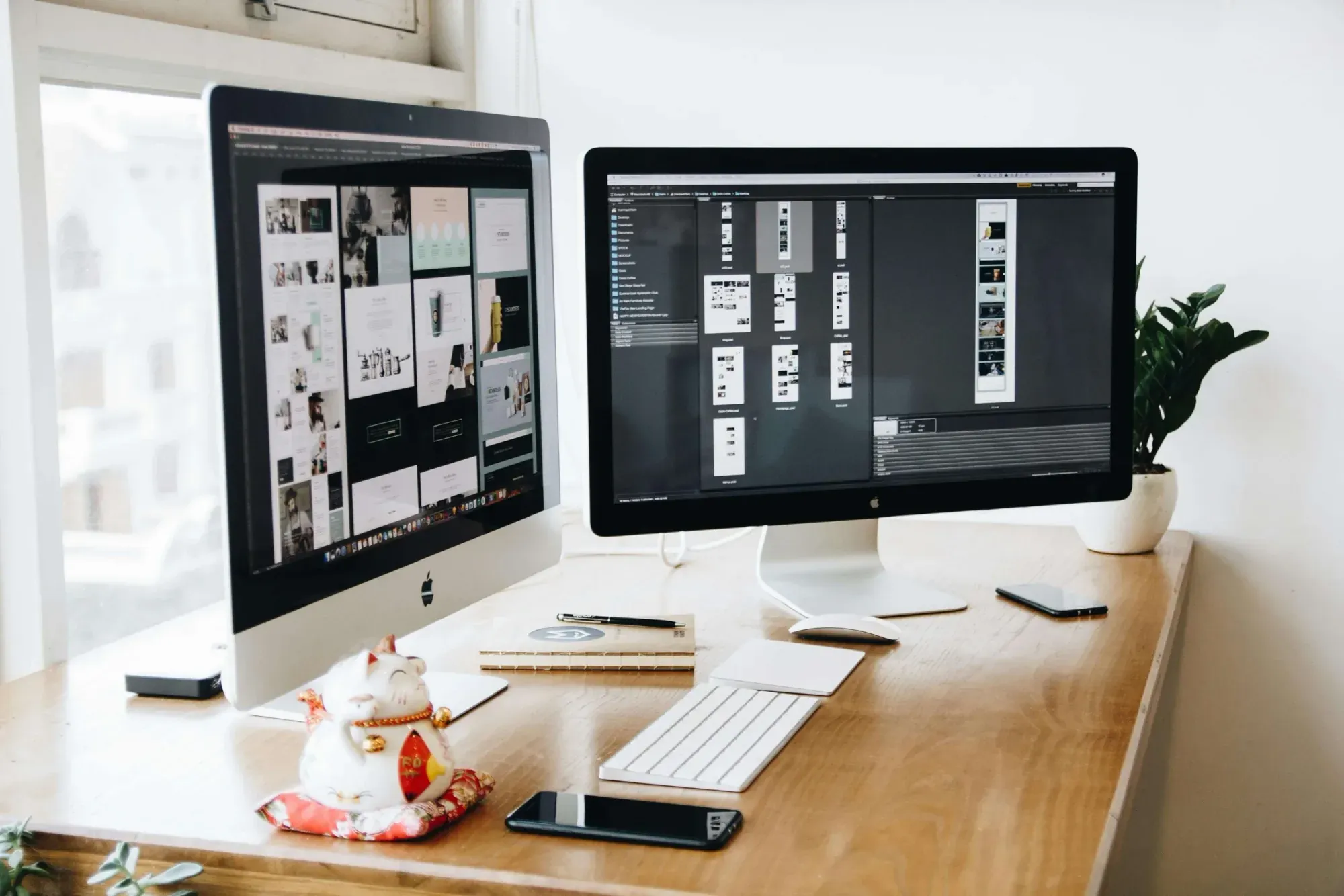 A workspace with two iMacs on a wooden desk displaying design projects, with a keyboard, mouse, smartphone, notebook and plant nearby.