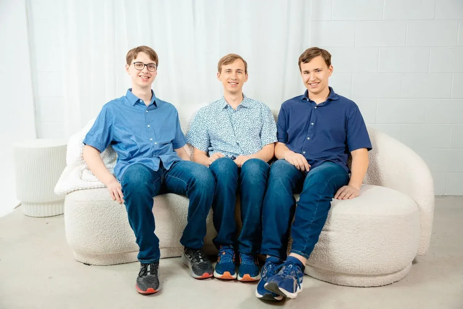 Three young men sitting on a light-coloured, plush couch in a bright room, smiling at the camera. They are dressed casually in blue shirts and jeans.