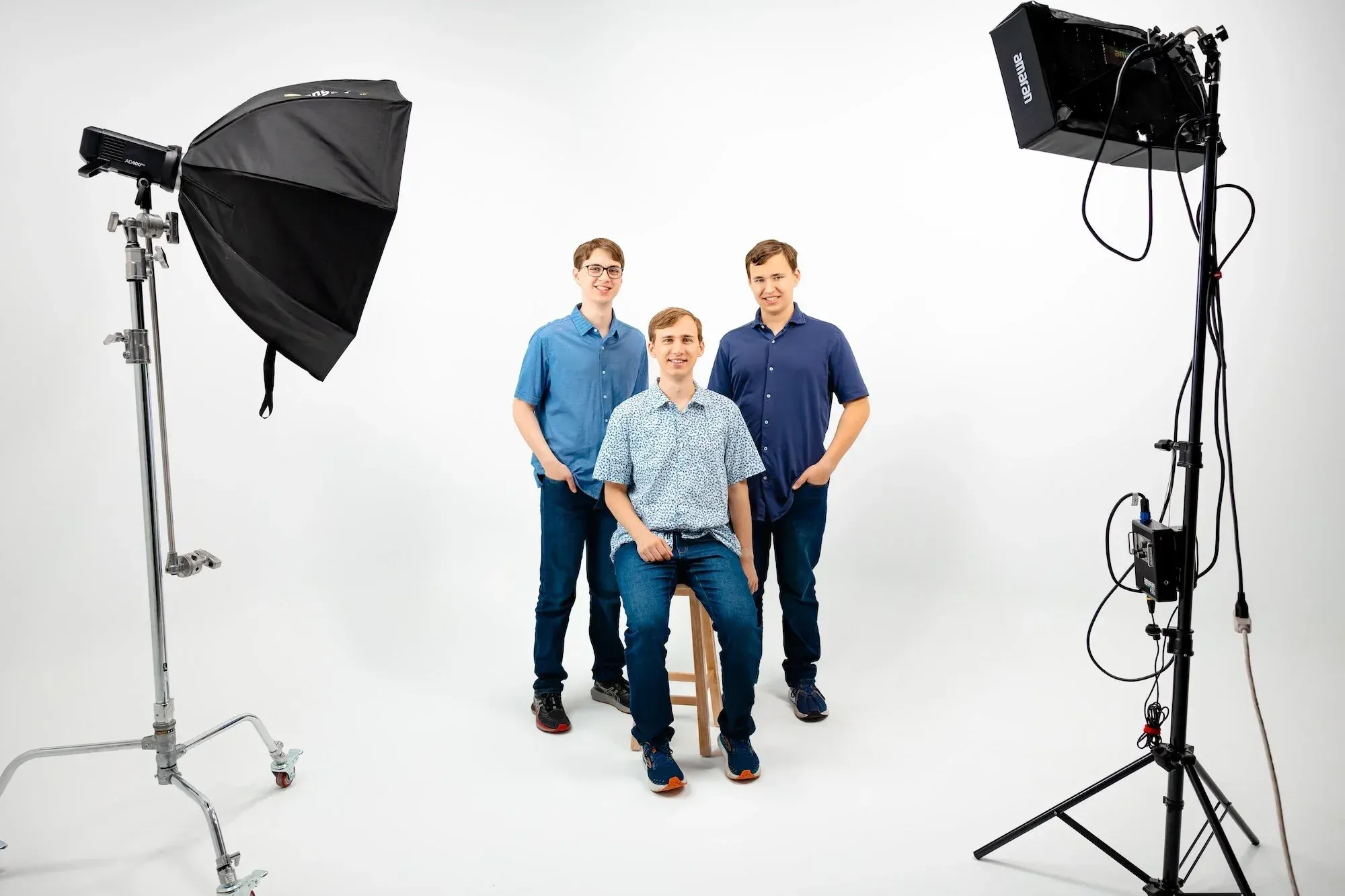 Three young men in casual clothing pose for a photo in a photography studio with professional lighting equipment and a plain white background.