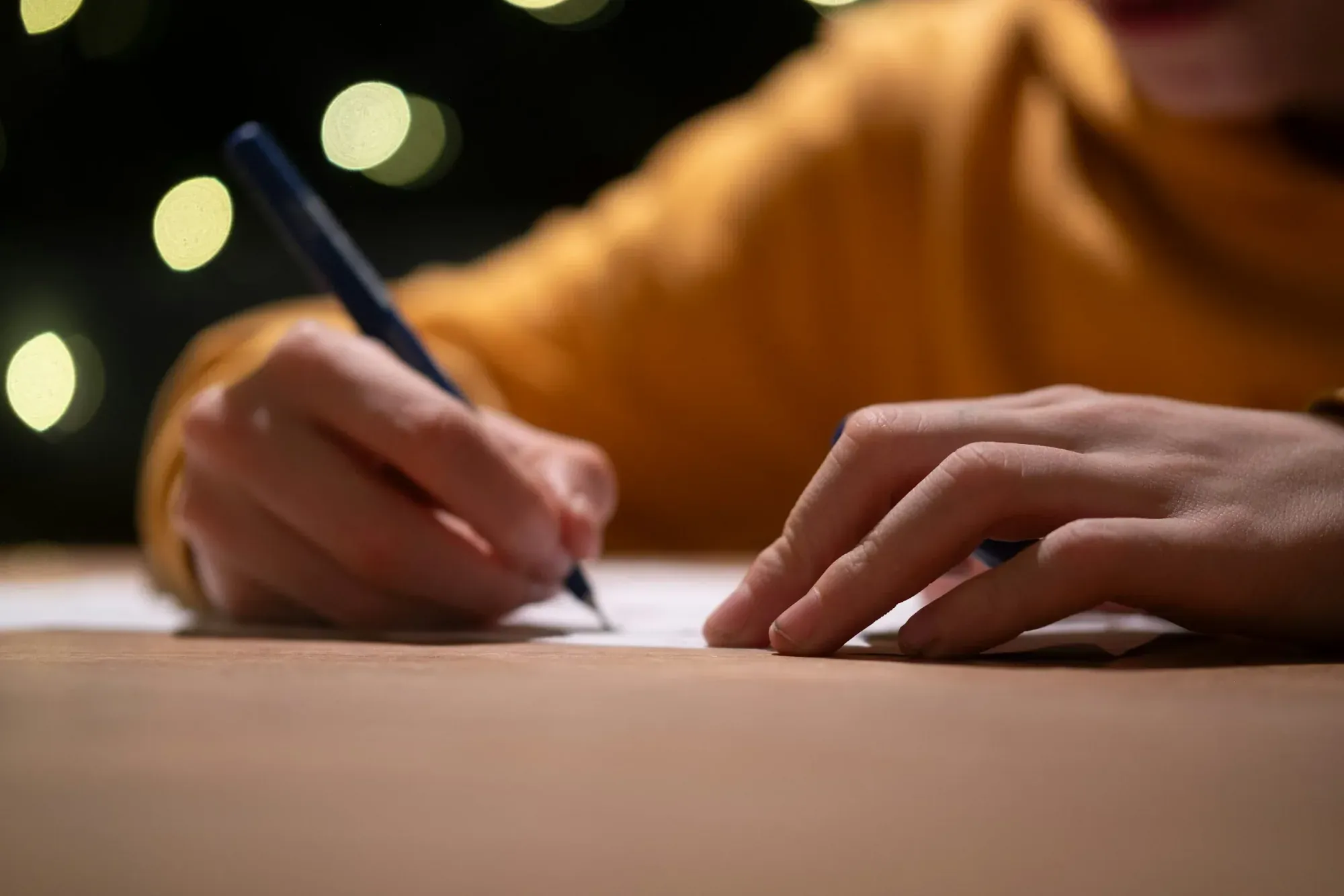 Close-up of a person writing with a pen on paper at night, blurred lights in the background.